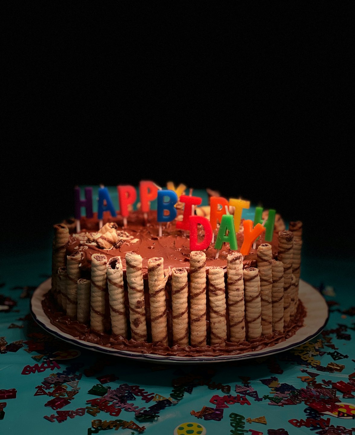 Chocolate birthday cake with wafer rolls and candles on turquoise tablecloth.